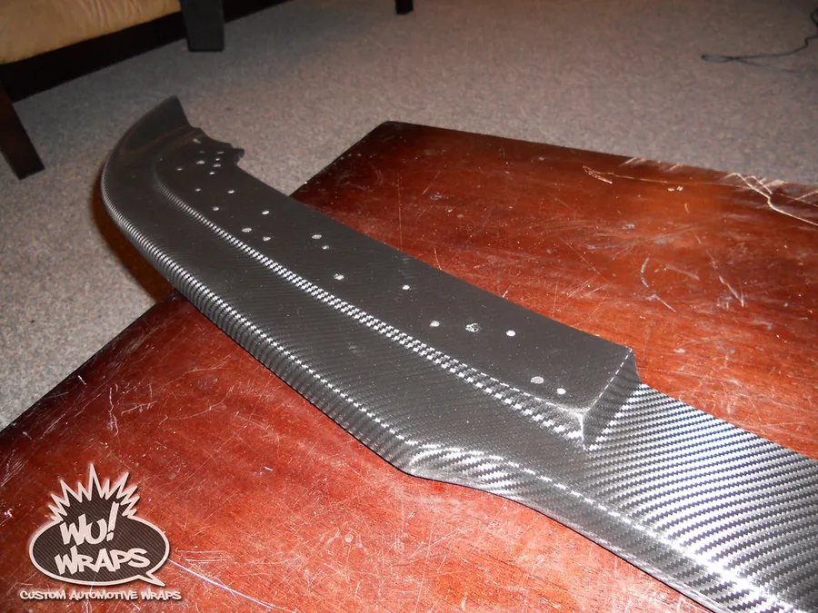 A finished carbon fiber wrapped front lip spoiler for a BMW E90, sitting on a wooden dining table