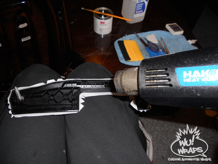 A BMW interior trim panel part-wrapped in carbon fiber film, resting on Mark's knees while he works a heat gun along the edge